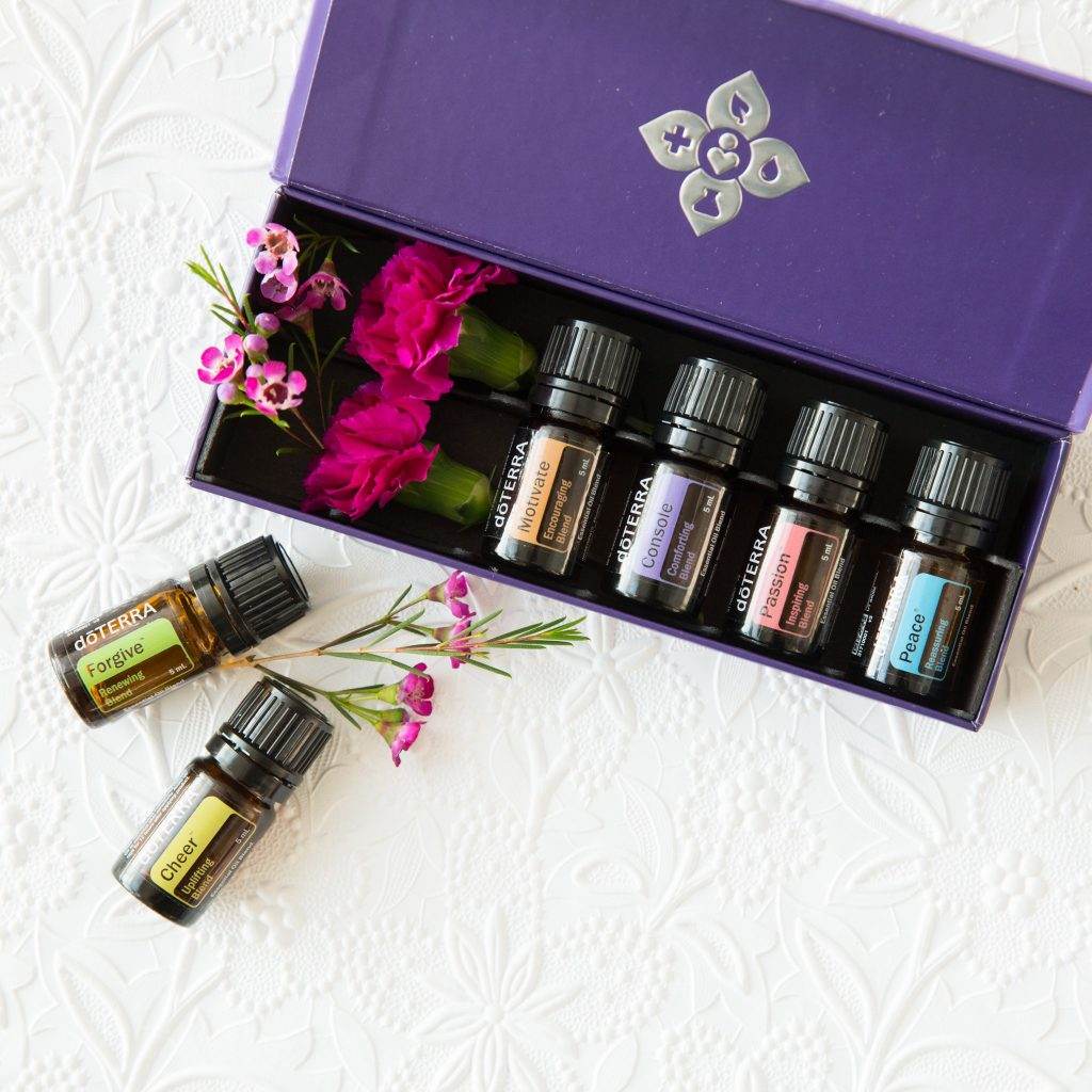 A purple box with floral design contains six essential oil bottles. Two additional bottles and small pink flowers are placed outside the box on a white textured surface.