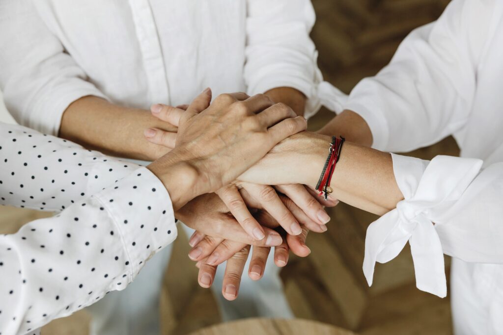 Four people place their hands together in a stack, symbolizing teamwork and unity at HolSol Wellness. They wear white shirts—one with black polka dots and one with a bracelet—reflecting a sense of collaboration and community.