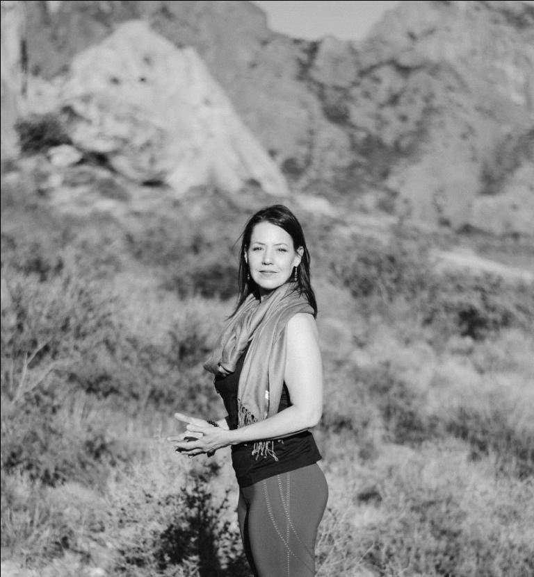 dvimukham profile landscape L Image woman with long dark hair wearing a CGM at sunset with wam welcoming look on her face. She's wearing a blue dress and holding an essential oil bottle