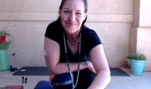White woman in her 40s with brown hair pulled back smiles at the camer after teaching a yoga class via Zoom. She is wearing black shirt and yoga pants and prayer beads made with blue stones. She is seated on a yoga block outside on front porch with a yoga mat behind her.