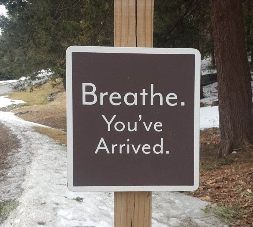 A sign on a wooden post reads "Breathe. You've Arrived." with snow and trees in the background.