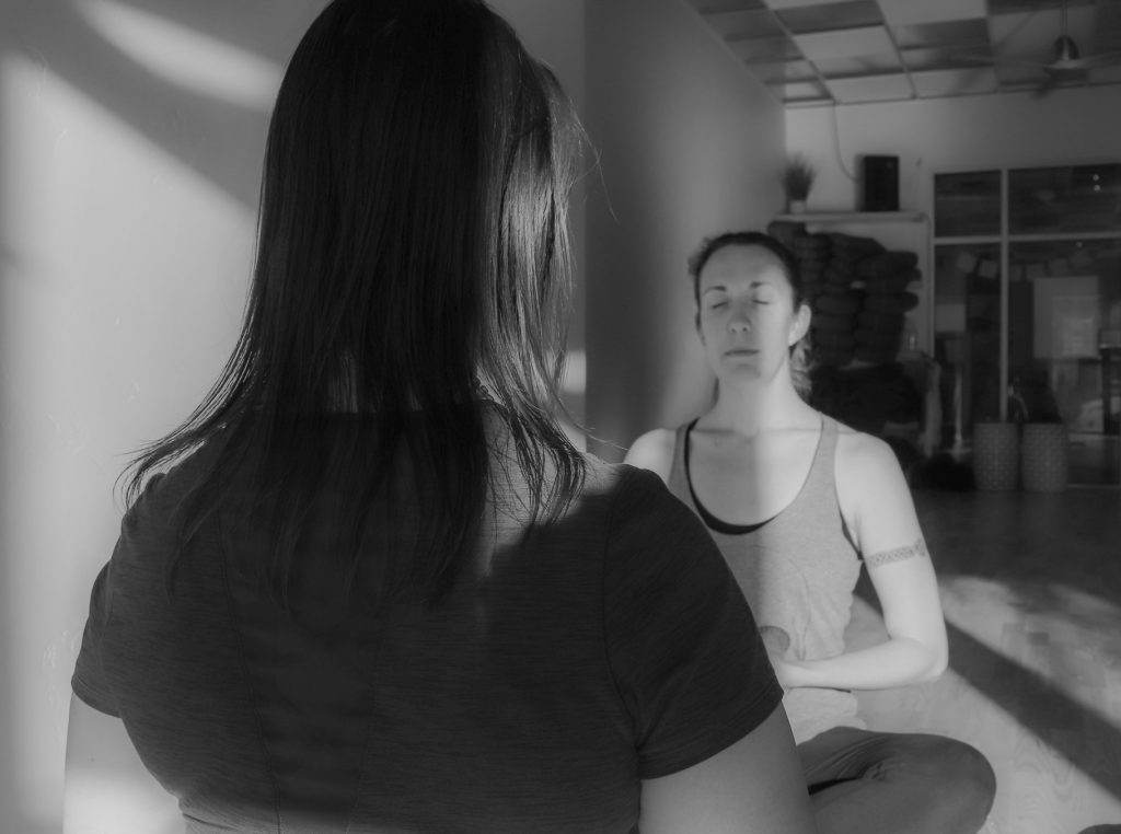 Two people face each other in a meditative posture inside a dimly lit room. One person is viewed from behind, while the other sits cross-legged with eyes closed, hands resting on their lap.