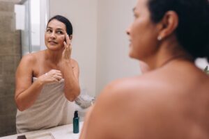 A person wrapped in a towel applies cream with geranium oil to their face while looking in the bathroom mirror. A small bottle is on the counter.