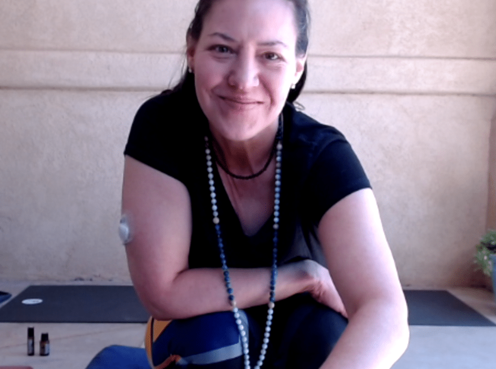 A person in a black shirt and a beaded necklace smiles while posing indoors. They have a medical device attached to their upper arm.
