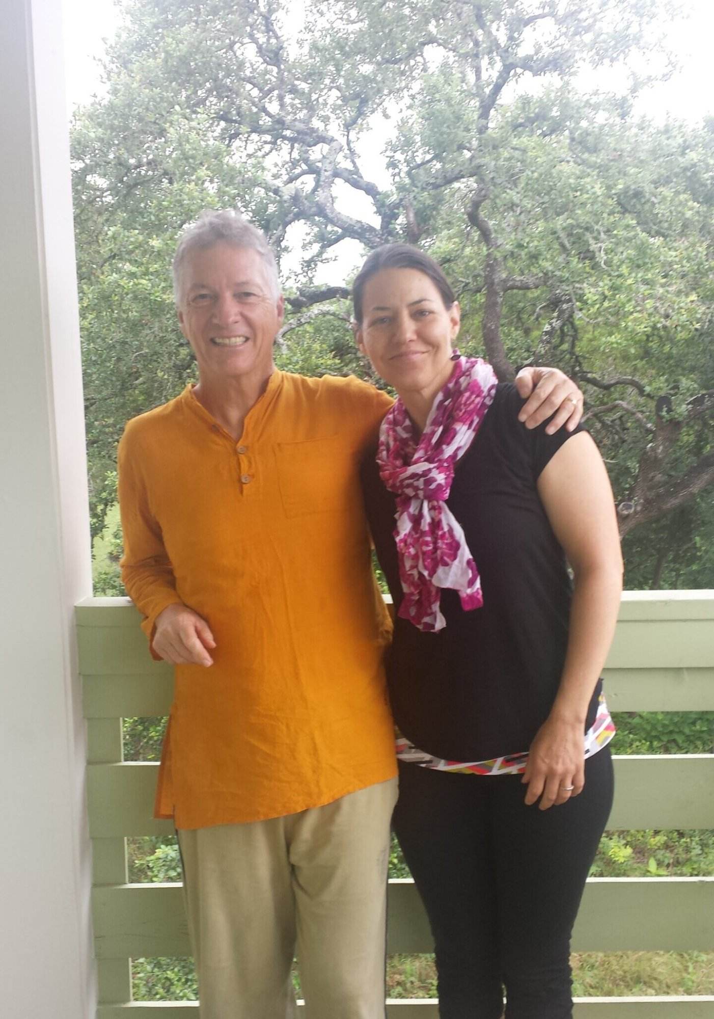 Dawn (the author) standing with her yoga teacher Joseph LePage on a second story porch with oak trees in the background. Dawn is wearing black yoga pants and top with a pink scarf. Joseph is weather tan pants and an orange button-down shirt