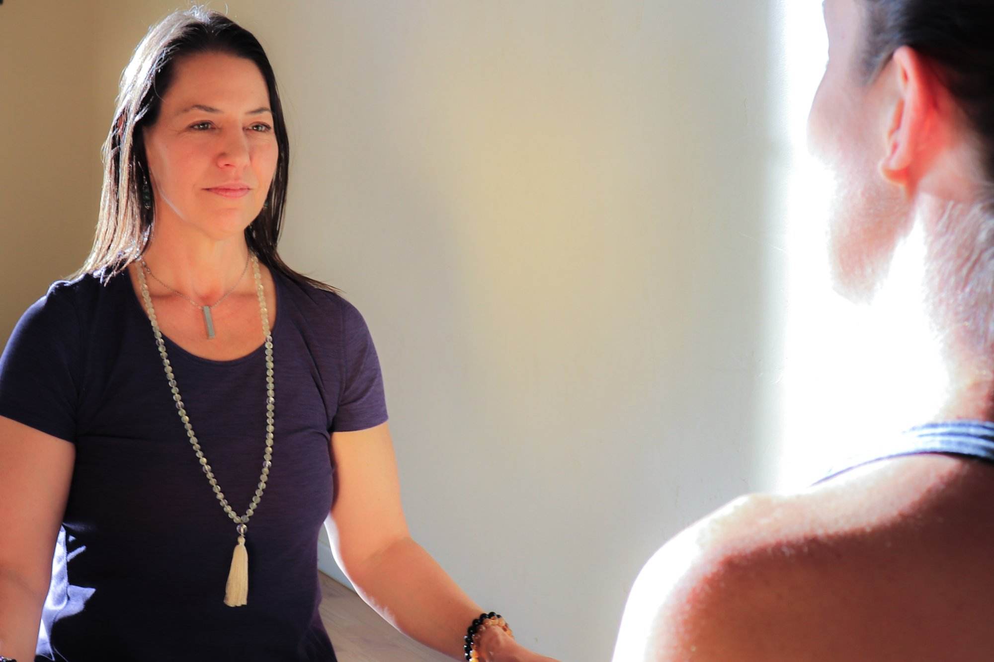 Yoga therapist, white woman in her 40s and shoulder-length brown hair observers her client, a white woman in her 30s as she notices her breath.