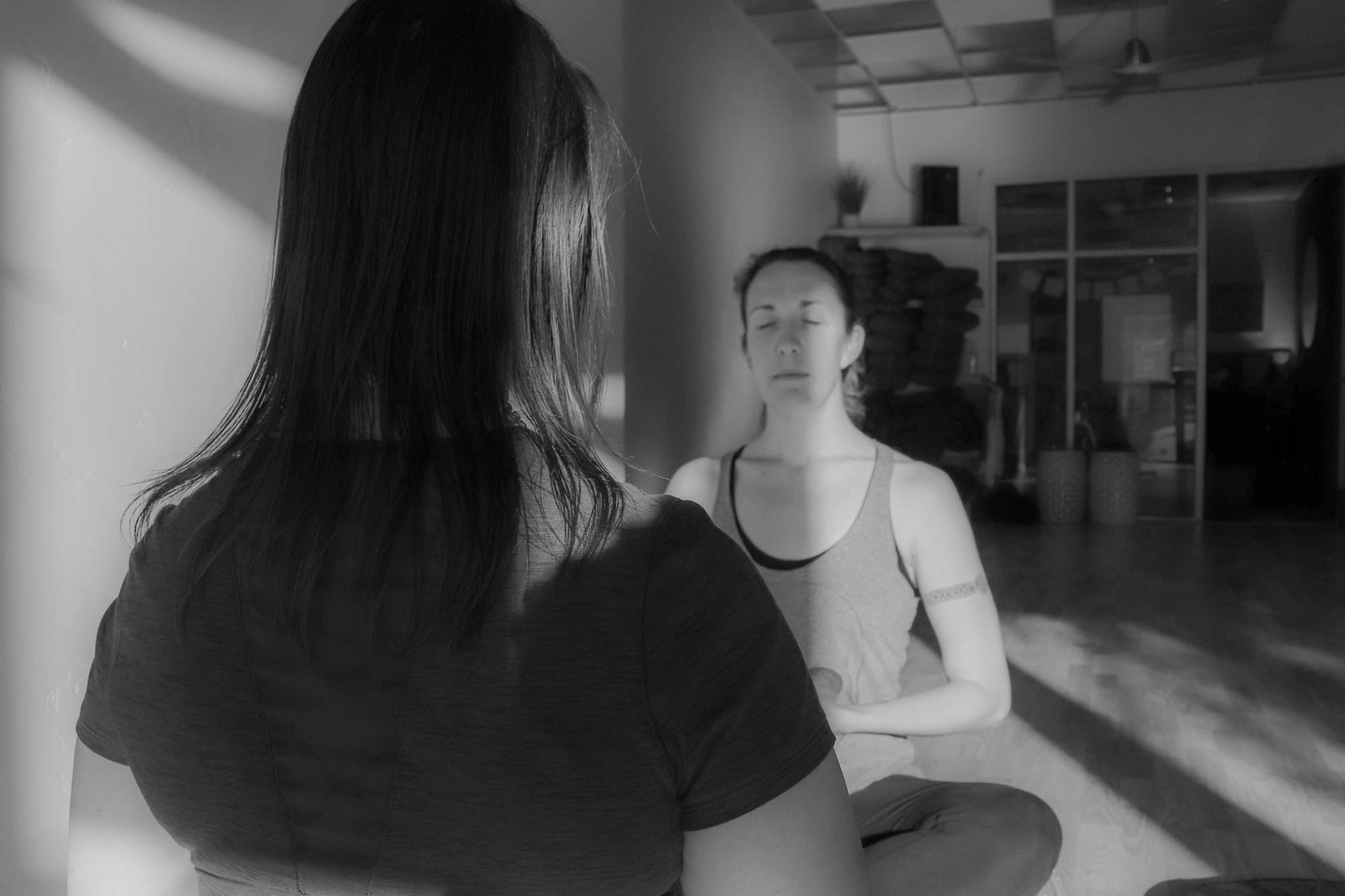 Black and white image of two white women with brown hair sitting in on wood floor of a yoga studio facing each other. Their eyes are closed at this point in the yoga therapy session.