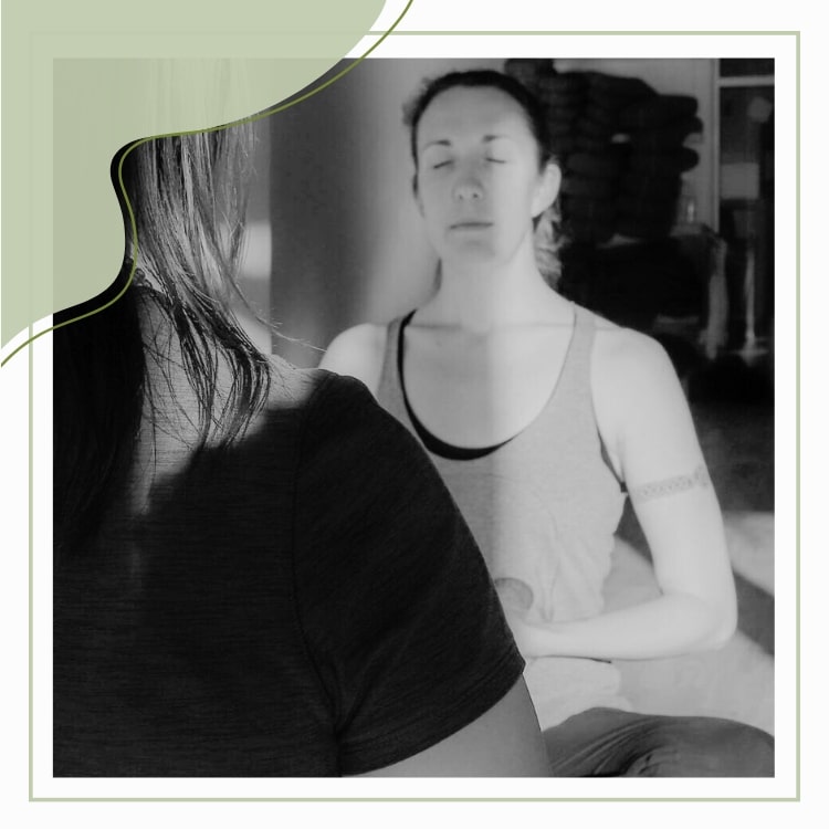 Black and white image of two white women with brown hair sitting in on wood floor of a yoga studio facing each other. Their eyes are closed at this point in the yoga therapy session.