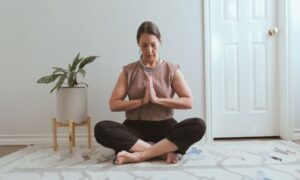 A woman sitting cross-legged on a rug, meditating with hands in prayer position, in a peaceful room with a plant beside her.