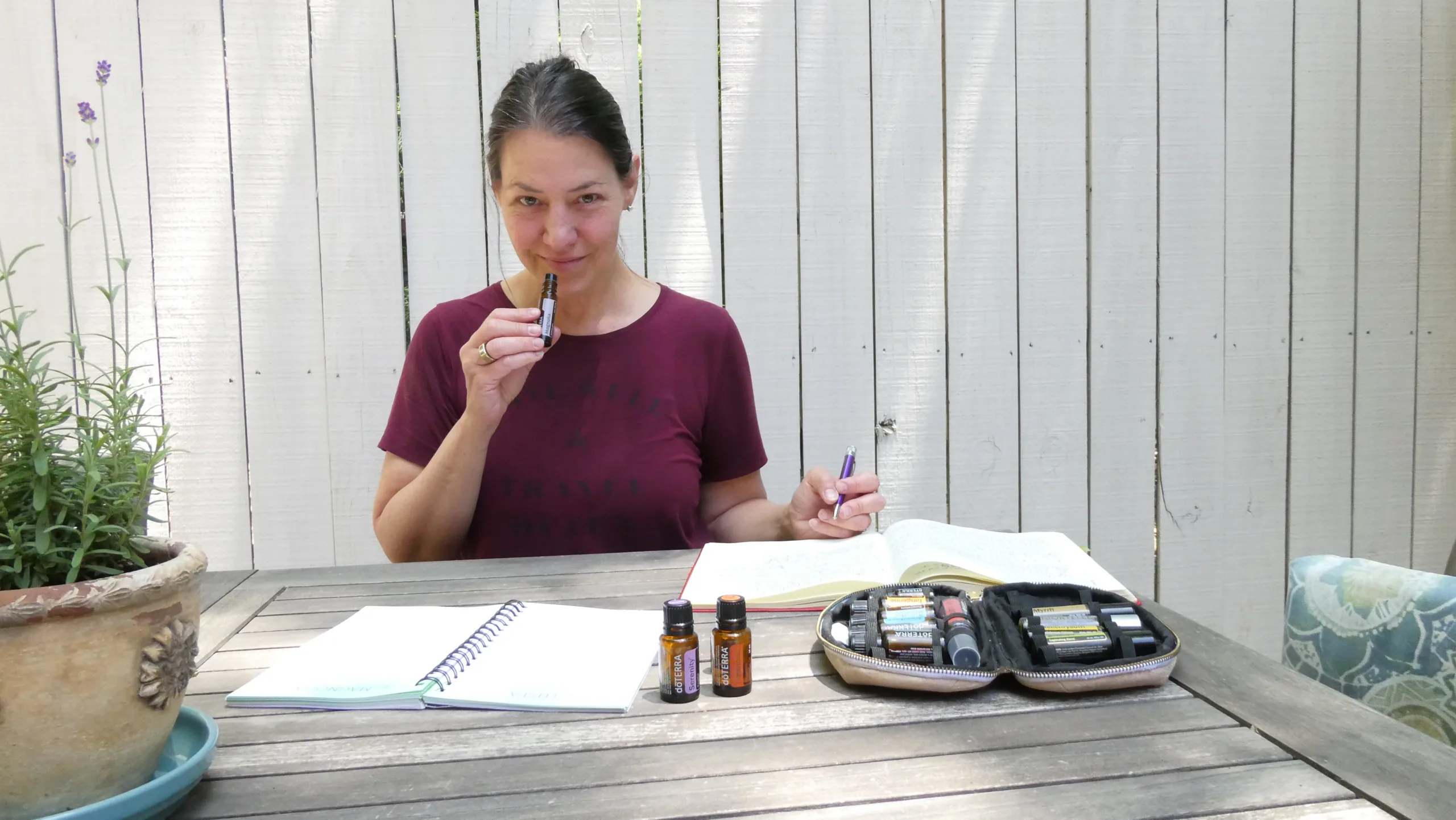 A person sits at a wooden table outside, holding a small vial near their nose. Nearby are notebooks, a pen, and several small bottles. A potted plant is on the table's left side.