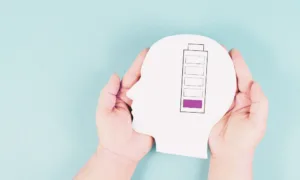 A person's hands hold a cutout of a human head with a battery gauge inside, highlighting the need to identify and overcome 12 hidden energy drains as it indicates a low charge level.