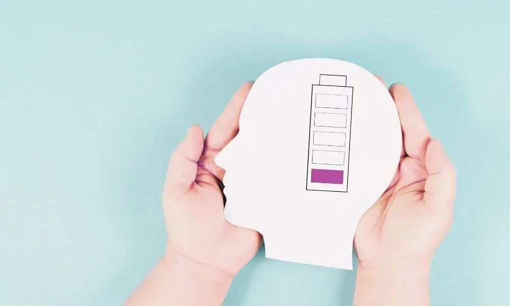 A person's hands hold a cutout of a human head with a battery gauge inside, highlighting the need to identify and overcome 12 hidden energy drains as it indicates a low charge level.