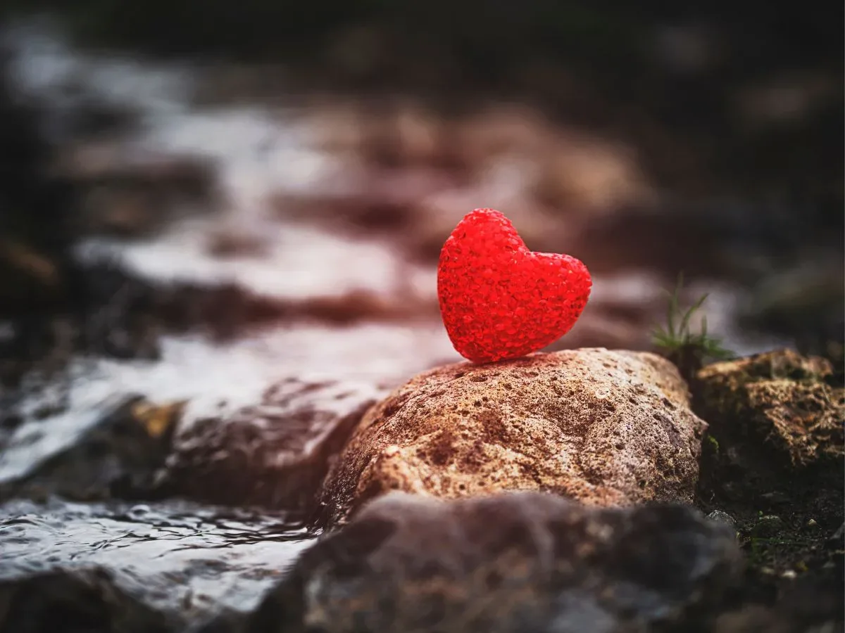 A textured red heart, symbolizing the process to mend a tender heart, rests on a rock by a flowing stream.