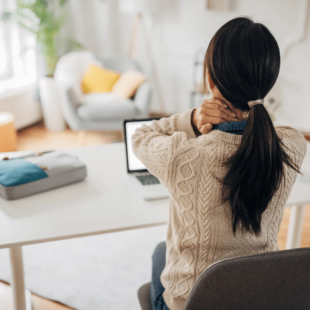 A person with long hair in a ponytail sits at a desk, using a laptop and rubbing their neck, suggesting discomfort or tension—perhaps seeking the soothing touch of a Neck Relief Aromatherapy Blend for Comfort.