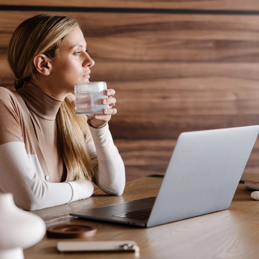Woman with a ponytail sits at a table, holding a glass, looking at a laptop. She is in a room with wooden walls and other items on the table.