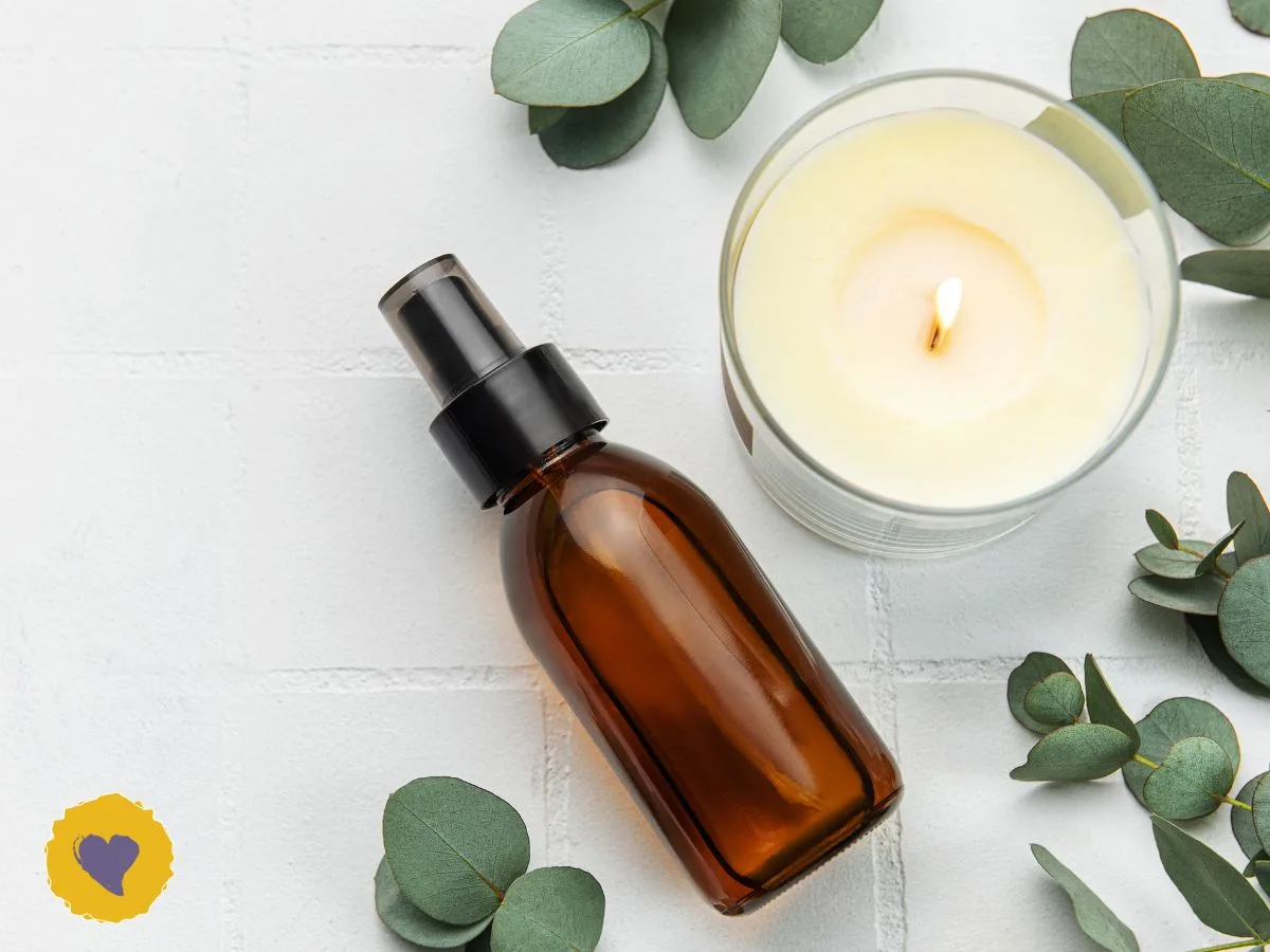 Eucalyptus Essential Oil: Nature’s Clean, Clarifying Ally - A brown glass spray bottle and a lit candle are arranged on a white surface. Eucalyptus leaves are scattered around.