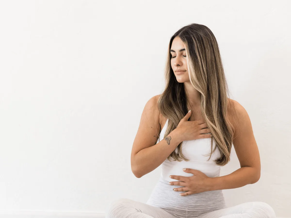Woman sits cross-legged against a white background, eyes closed, with one hand on her chest and the other on her abdomen, appearing to practice deep breathing or meditation.