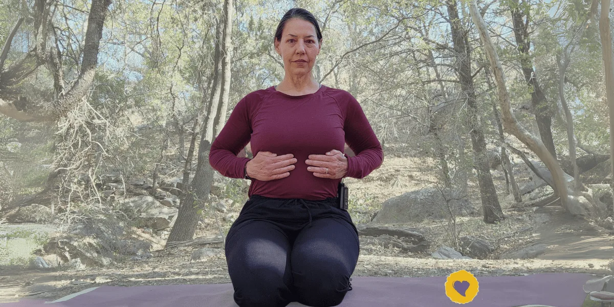 Blog2 Adham Sparsha Mudra-min A woman sits on a yoga mat outdoors, kneeling with her hands on her abdomen, practicing a breathing exercise in a forest setting.