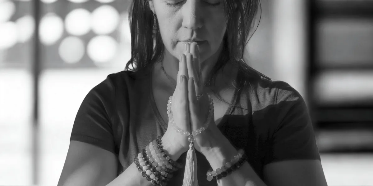 A woman with prayer beads on her wrists holds her hands in a prayer position near her face, appearing to meditate or pray, possibly practicing breathing for anxiety.