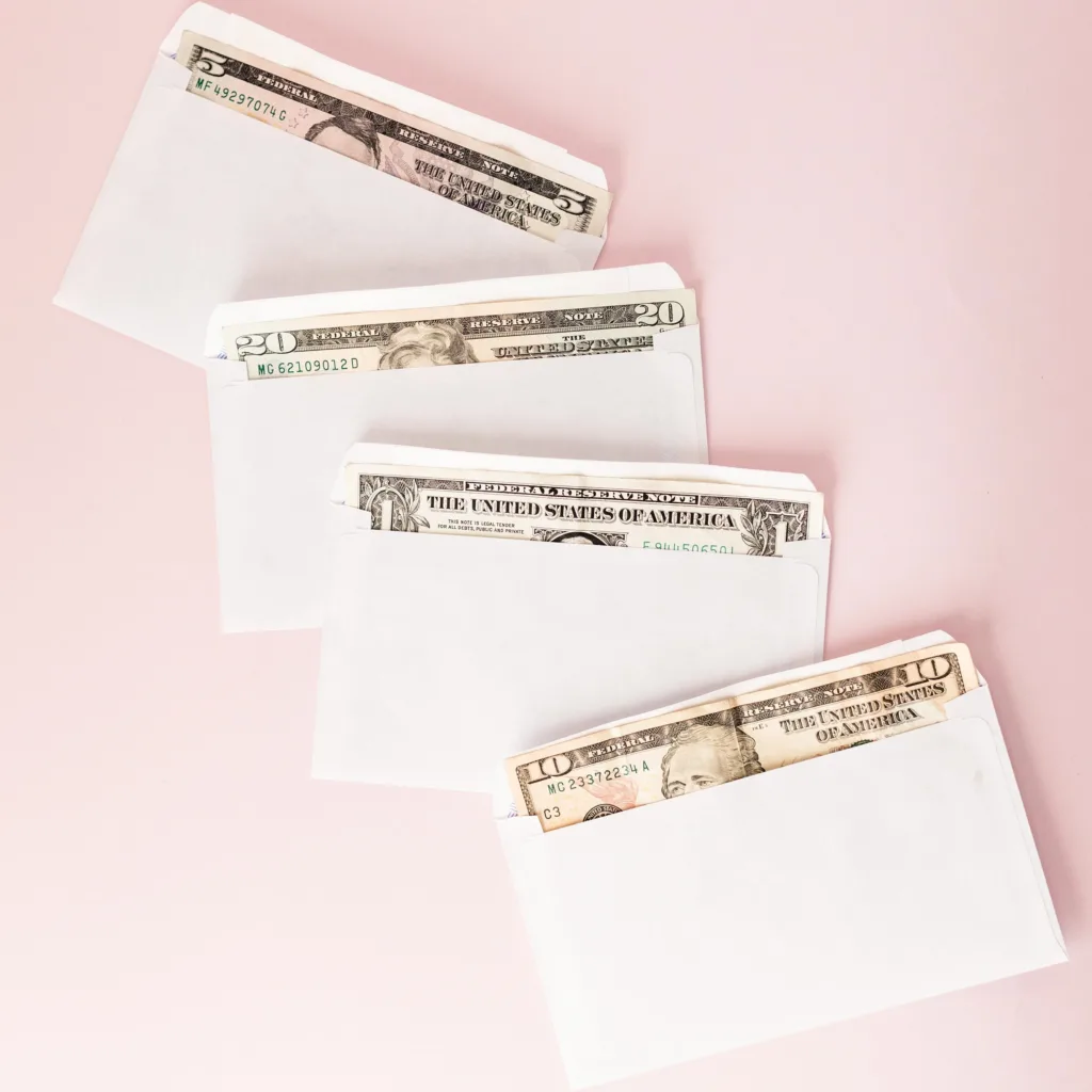 Budget money envelopes Four white envelopes, each containing U.S. paper currency in different denominations, are arranged diagonally on a light pink background.