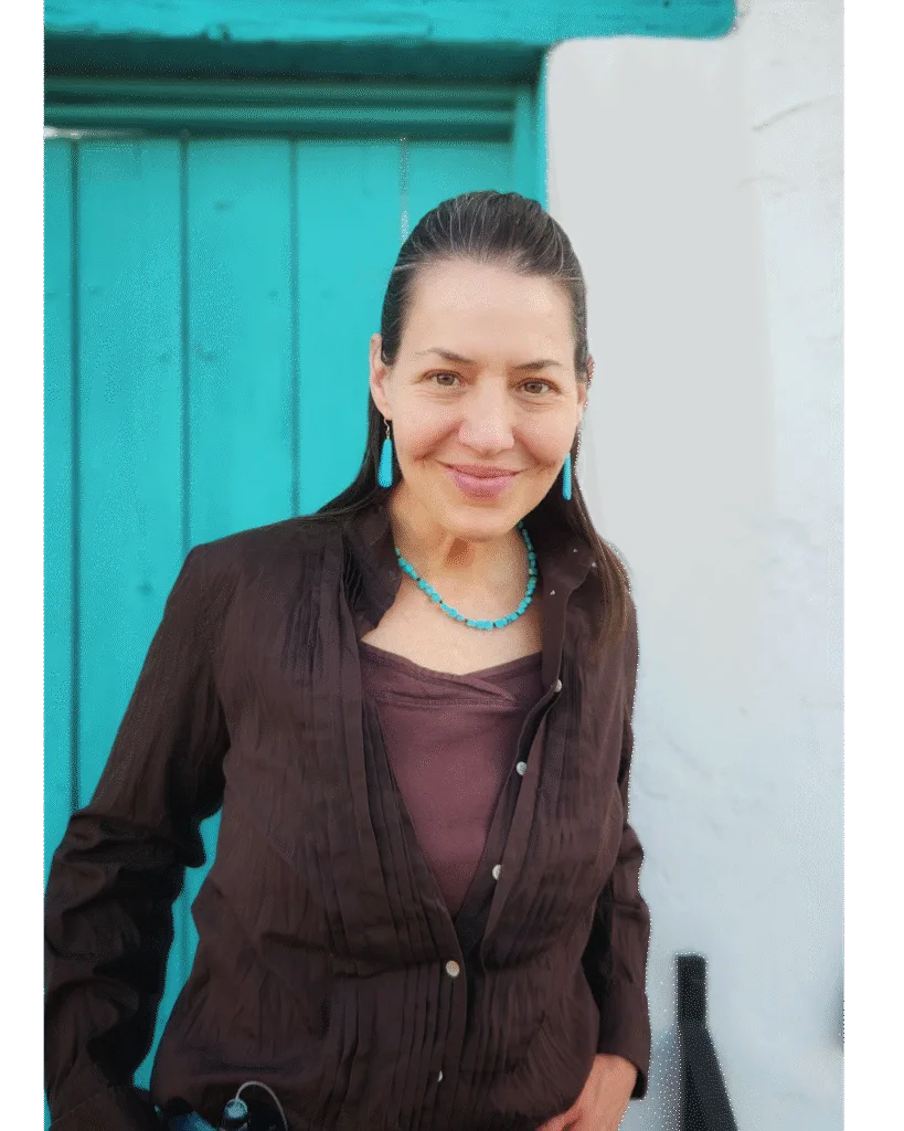 A woman with long brown hair, wearing a brown shirt, turquoise necklace, and earrings, stands smiling in front of a turquoise door and white wall.