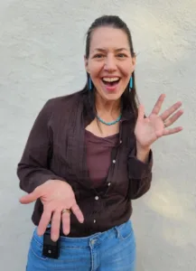 A woman with long hair smiles and waves with both hands, wearing a brown shirt, blue jeans, and turquoise jewelry, standing against a light-colored wall—radiating the calm confidence of somatic alchemy yoga therapy.