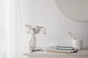 A white desk with a vase of dried flowers, a pair of glasses, a tablet, notebooks, and a cup holding pencils, set against a textured white wall with part of a round mirror visible.