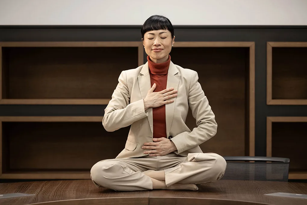 Woman in suit Hasta mudras on desk-min_1020x680 A woman in a beige suit sits cross-legged on a table with eyes closed, one hand on her chest and one on her abdomen, appearing to practice mindful breathing.