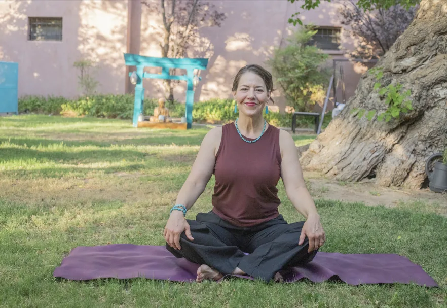 A woman sits cross-legged on a purple yoga mat outdoors, smiling, with trees and a building in the background.