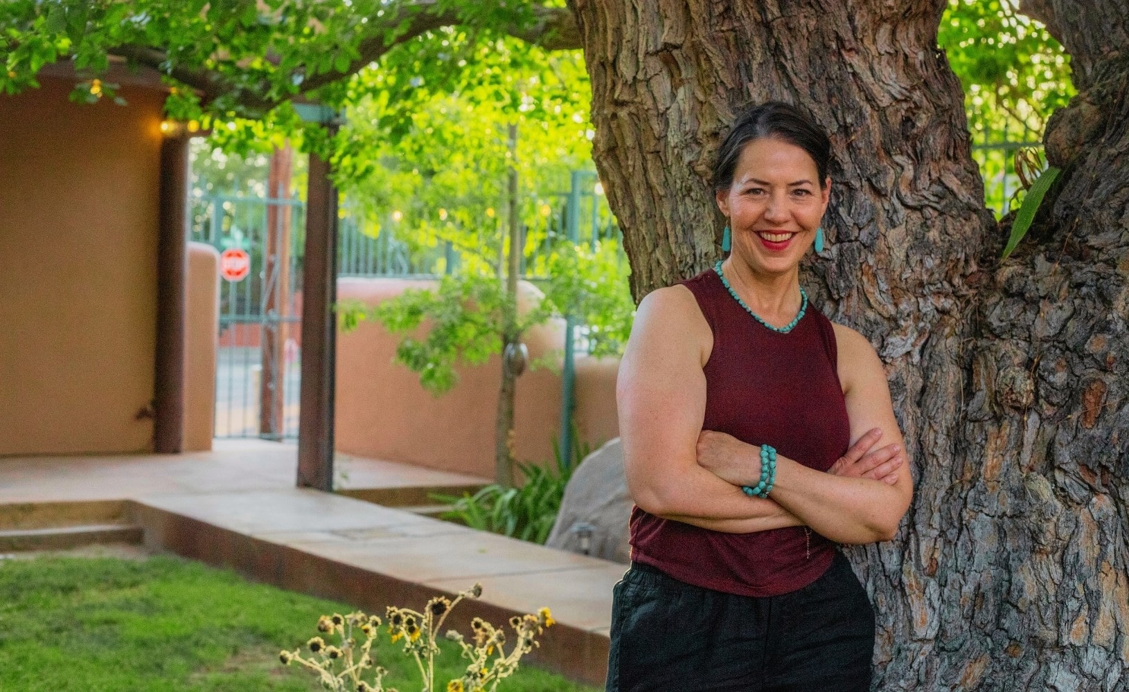 A woman stands with arms crossed, leaning against a large tree in a garden, with a building and greenery visible in the background.