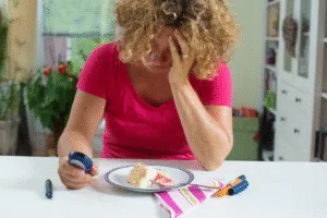 A person in a pink shirt sits at a table, looking concerned at a glucose meter and a plate with cake, highlighting the importance of stress management for those living with diabetes, with insulin supplies nearby.