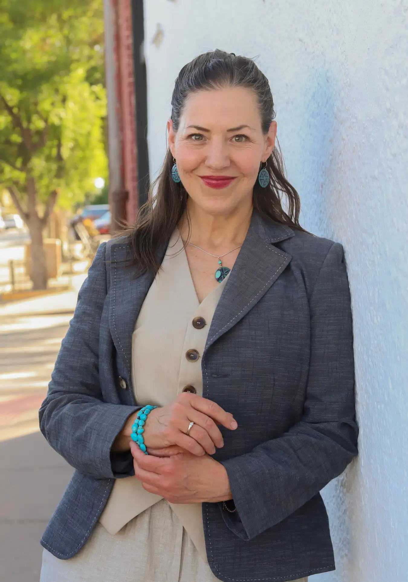 A woman with long dark hair, wearing a gray blazer and beige outfit, stands outdoors against a white wall, smiling at the camera. She is accessorized with turquoise jewelry.