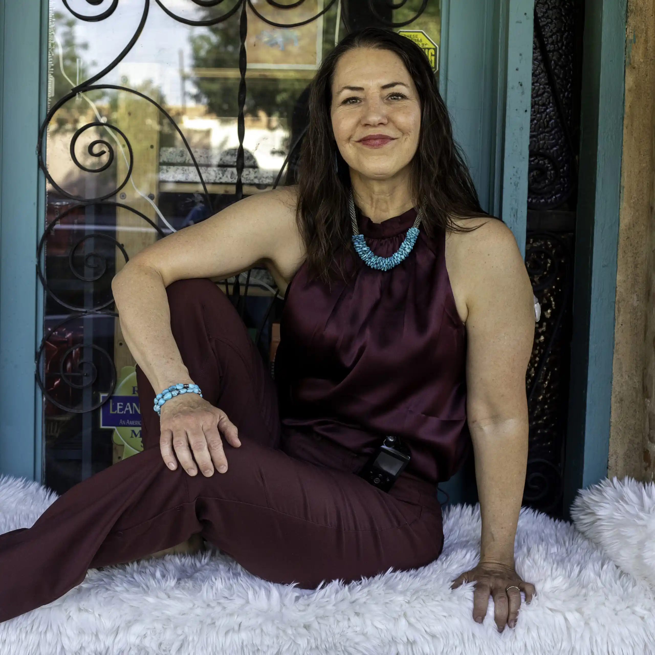 A woman with long brown hair, wearing a sleeveless maroon top and pants, sits on a white furry cushion in front of a decorative door, smiling at the camera.