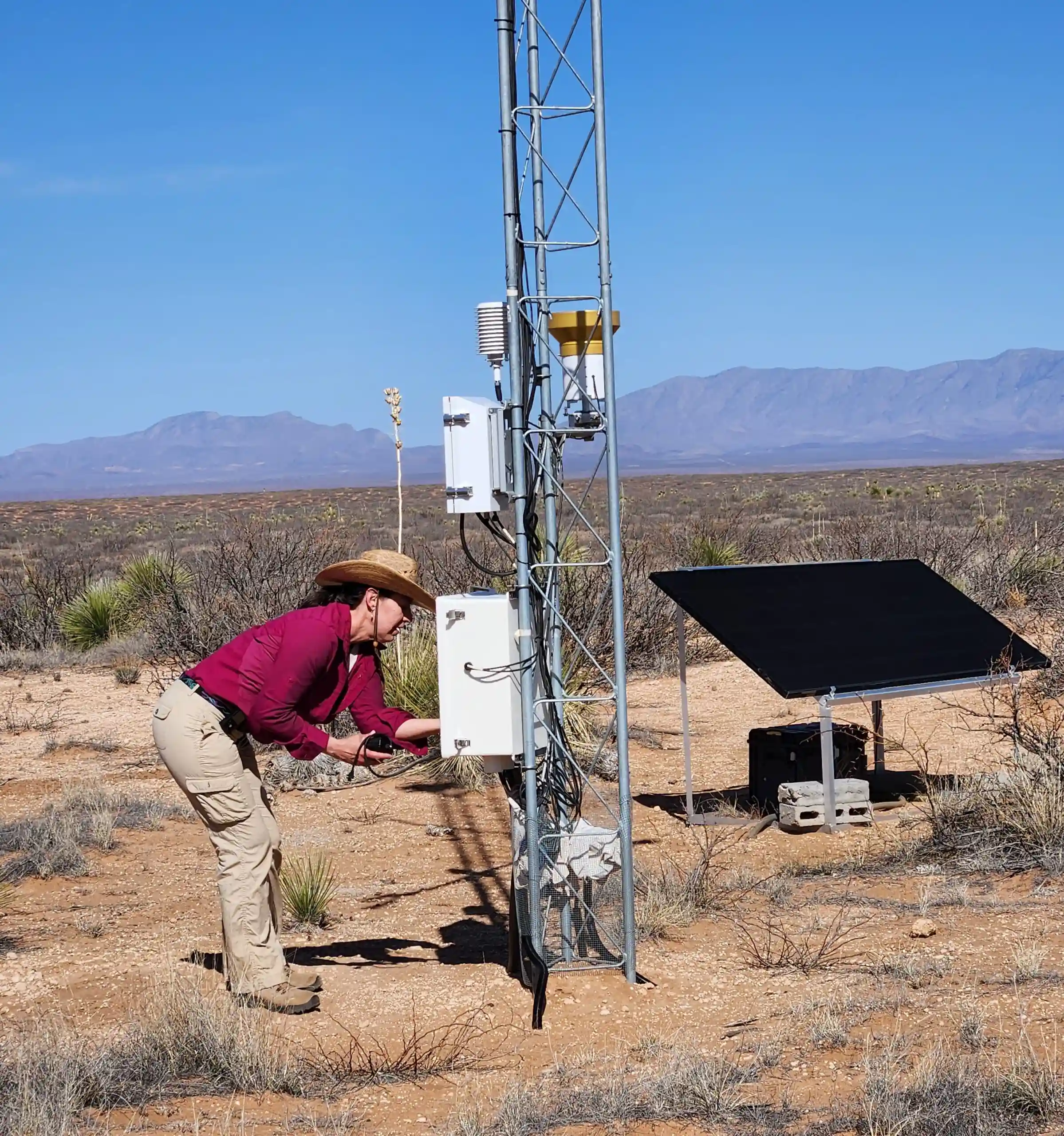 A person in outdoor clothing works on weather monitoring equipment at a metal tower in a desert landscape with mountains in the background and a solar panel nearby.