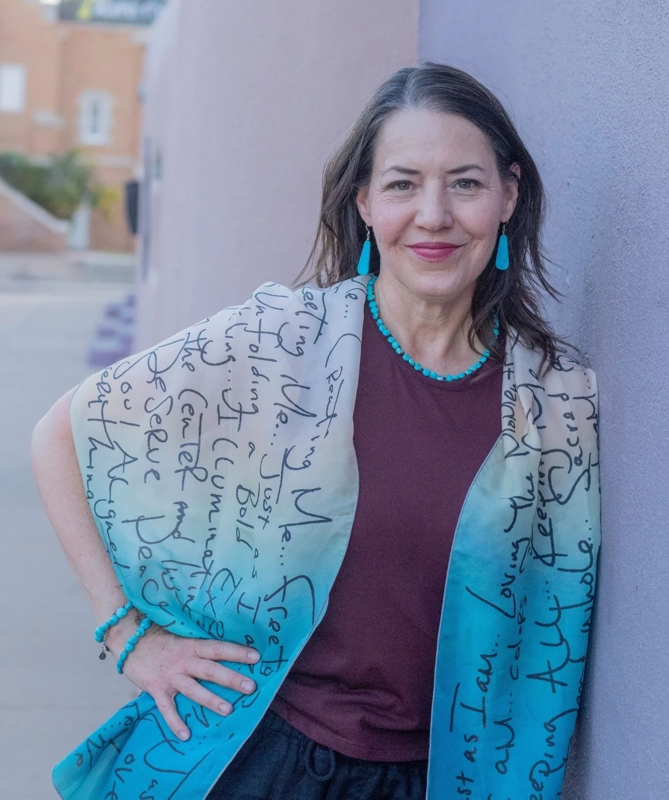 Woman with long brown hair, wearing turquoise jewelry and a blue shawl with script writing, stands against a lavender wall, smiling with one hand on her hip.