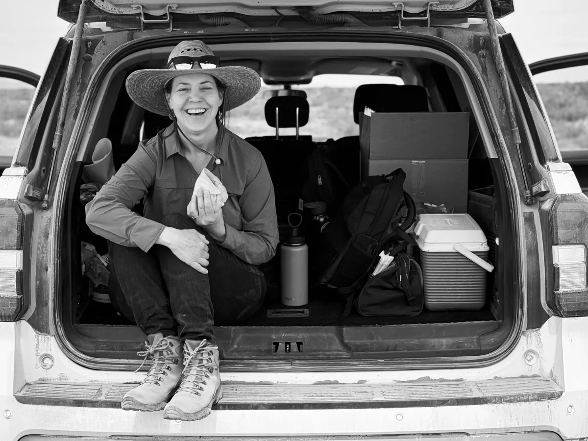 Dawn Browning wearing a wide-brimmed hat sits smiling in the open trunk of an SUV, surrounded by gear, a cooler, boxes, and Essential Oils: Your Path to Holistic Health.