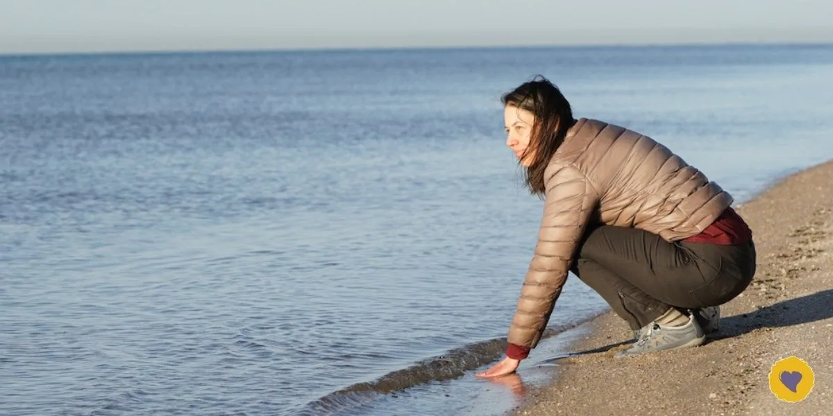 Dawn Browning in a brown jacket squats at the edge of a calm beach, touching the water with her hand under a clear sky, embodying a moment of mindfulness—an example of how to manage your energy by connecting with nature.