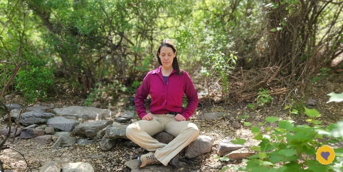 Dawn Browning sits cross-legged on rocks in a sunlit wooded area, eyes closed, wearing a magenta jacket and beige pants—a serene moment that beautifully illustrates how to manage your energy amidst nature.